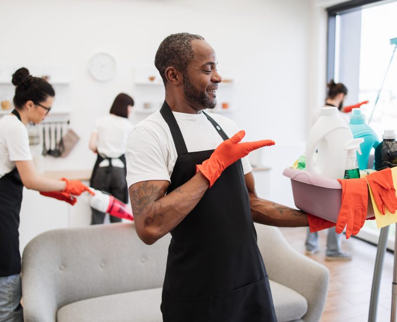 Portrait of young African American man professional cleaning worker pointing on bucket for washing with detergents on bright kitchen studio background, copy space.